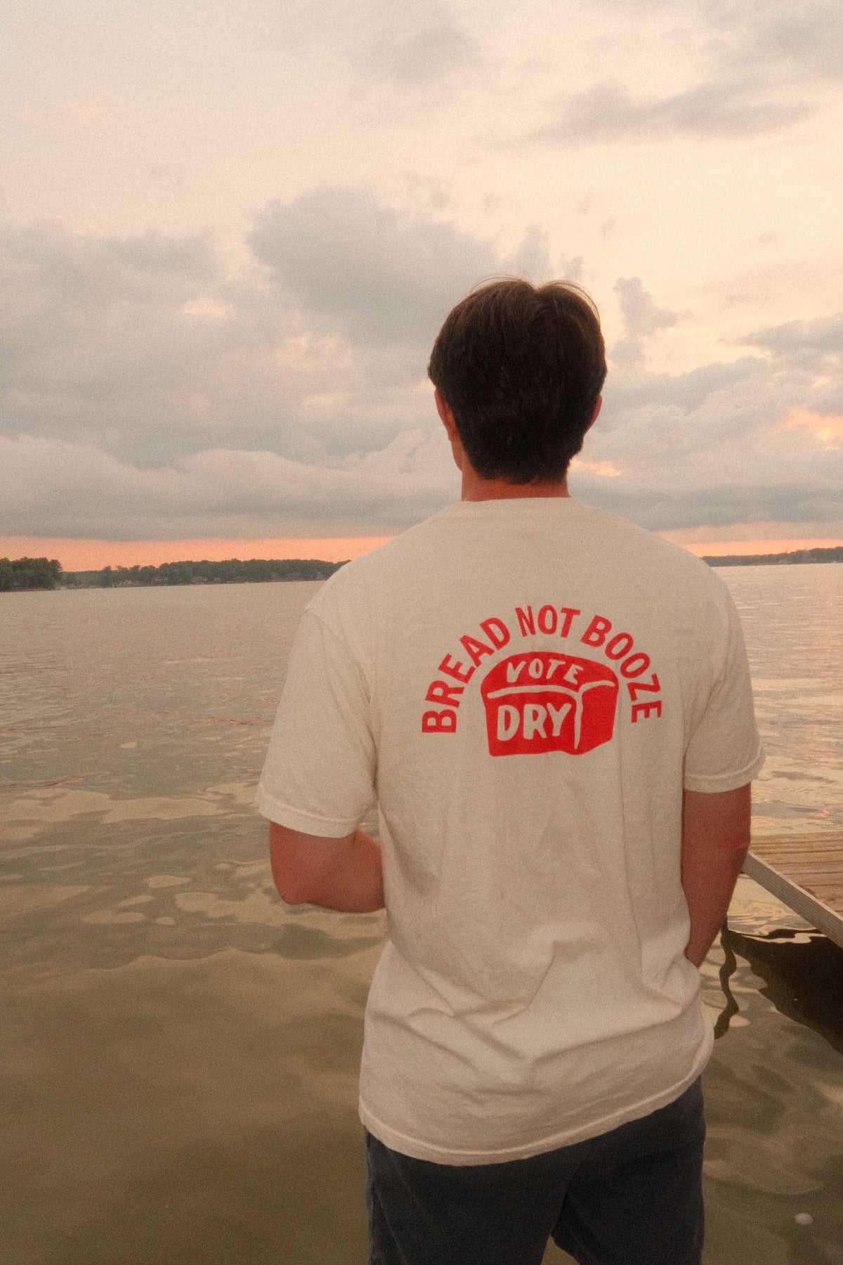 White Ohio Anti-Saloon League Bread Not Booze heavyweight t-shirt worn on a dock at sunset, back graphic reading Bread Not Booze Vote Dry