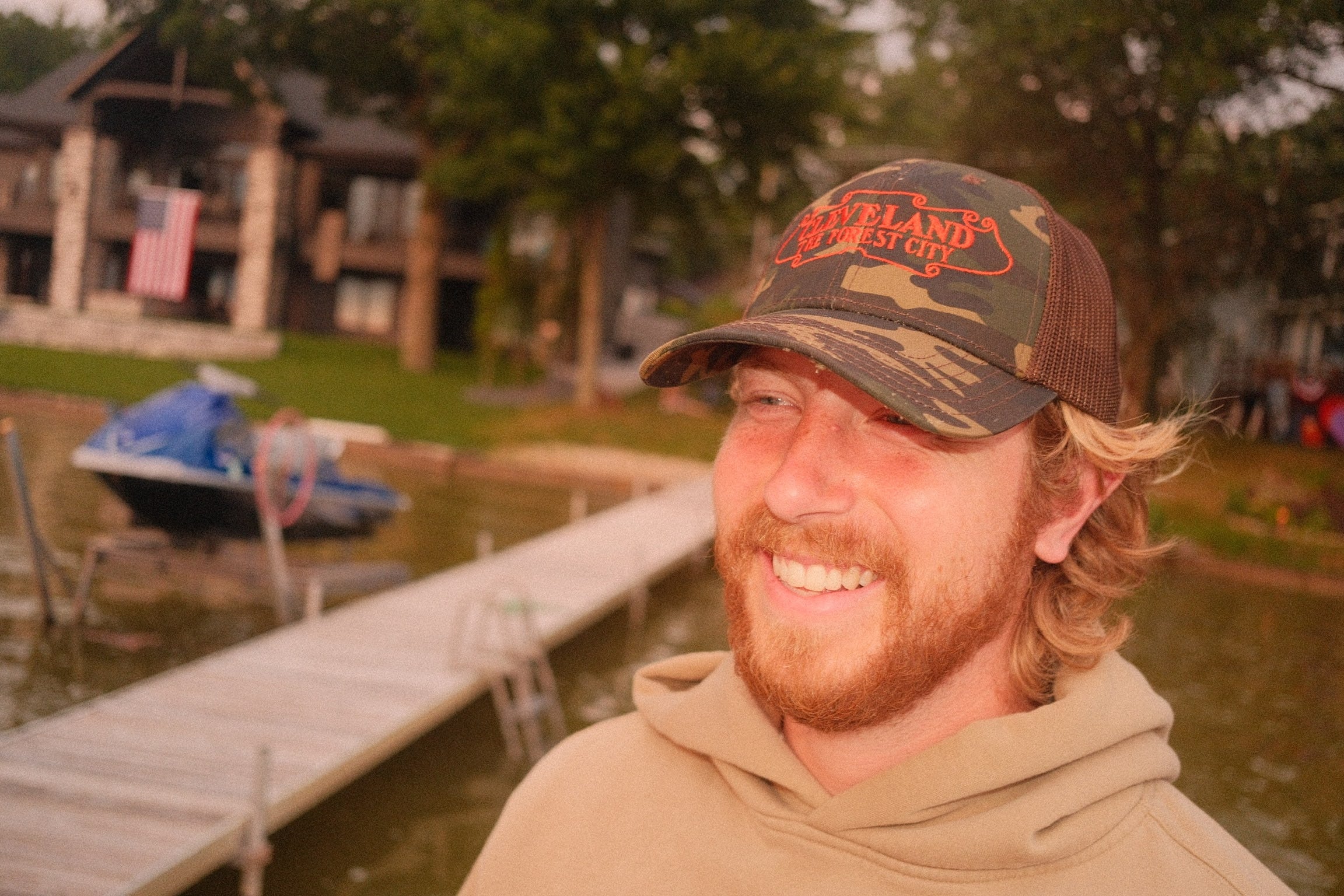 A smiling man with a beard and blonde hair wearing a camouflage trucker hat with "Cleveland The Forrest City" embroidered in Orange, standing outdoors near a dock and water.