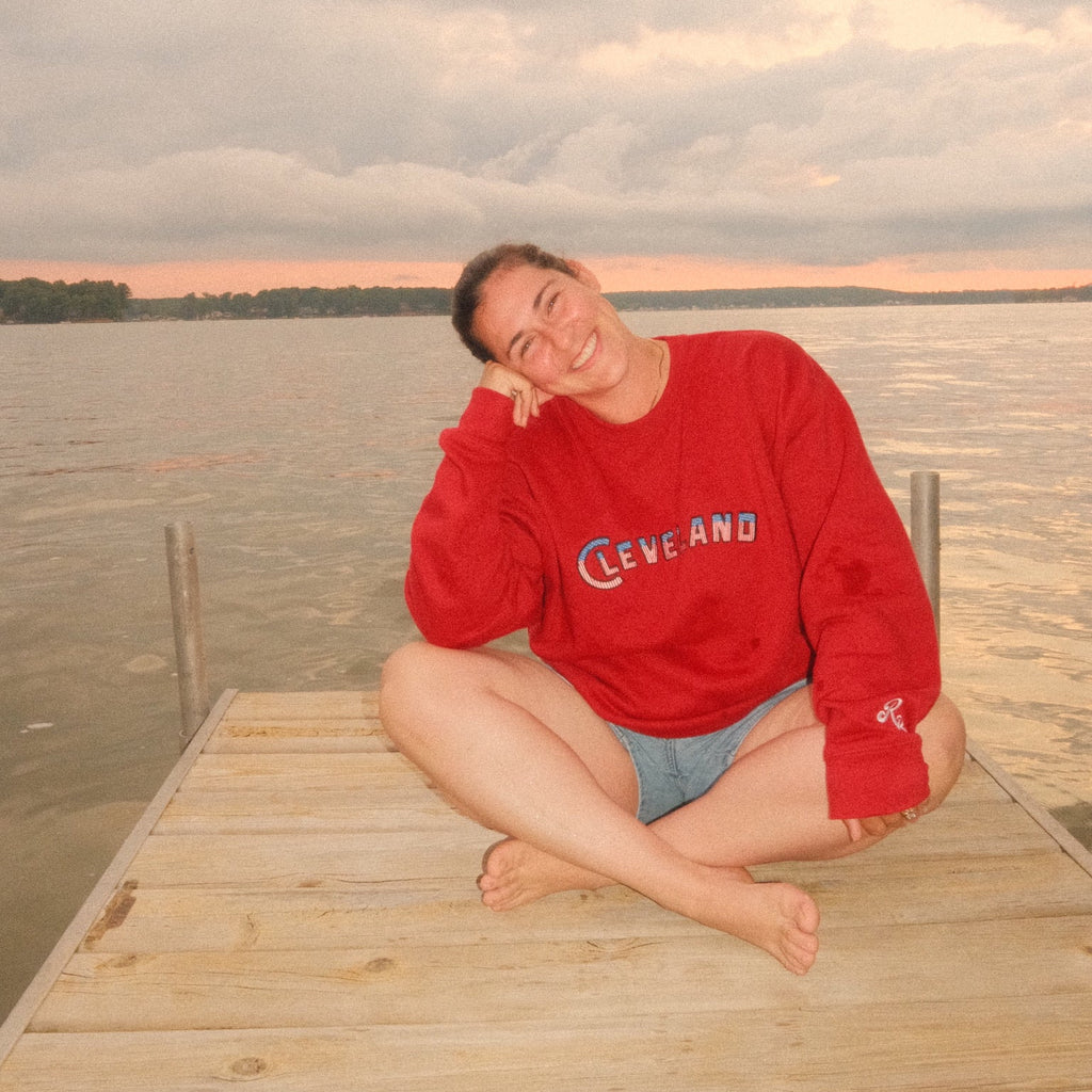 A person sitting cross-legged on a wooden dock by the water, wearing a red sweatshirt with 'CLEVELAND' printed on the front and light blue shorts, under an overcast sky.