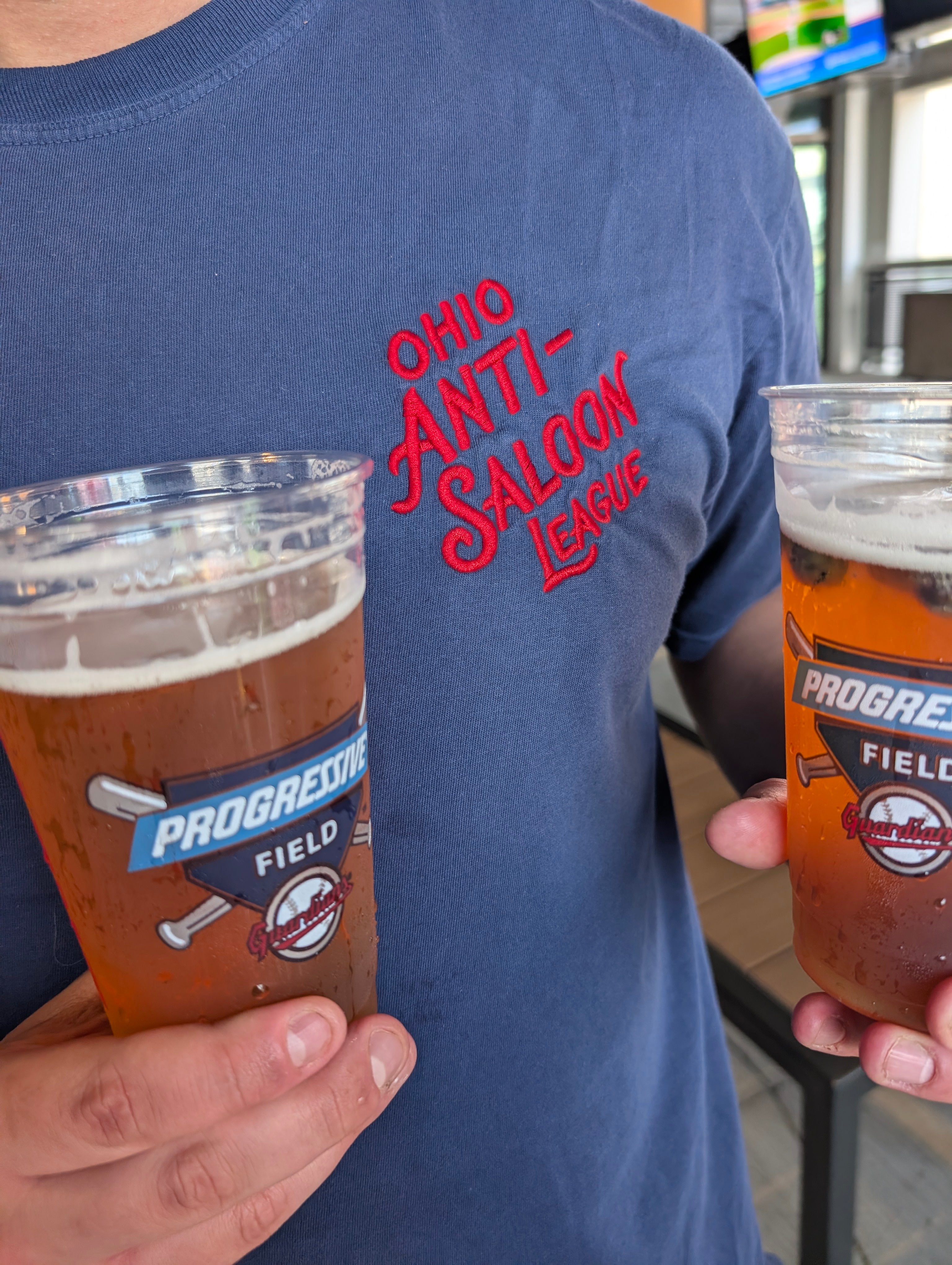 A person wearing a navy blue t-shirt with red 'Ohio Anti-Saloon League' text, holding two clear plastic cups of amber beer with foam heads.