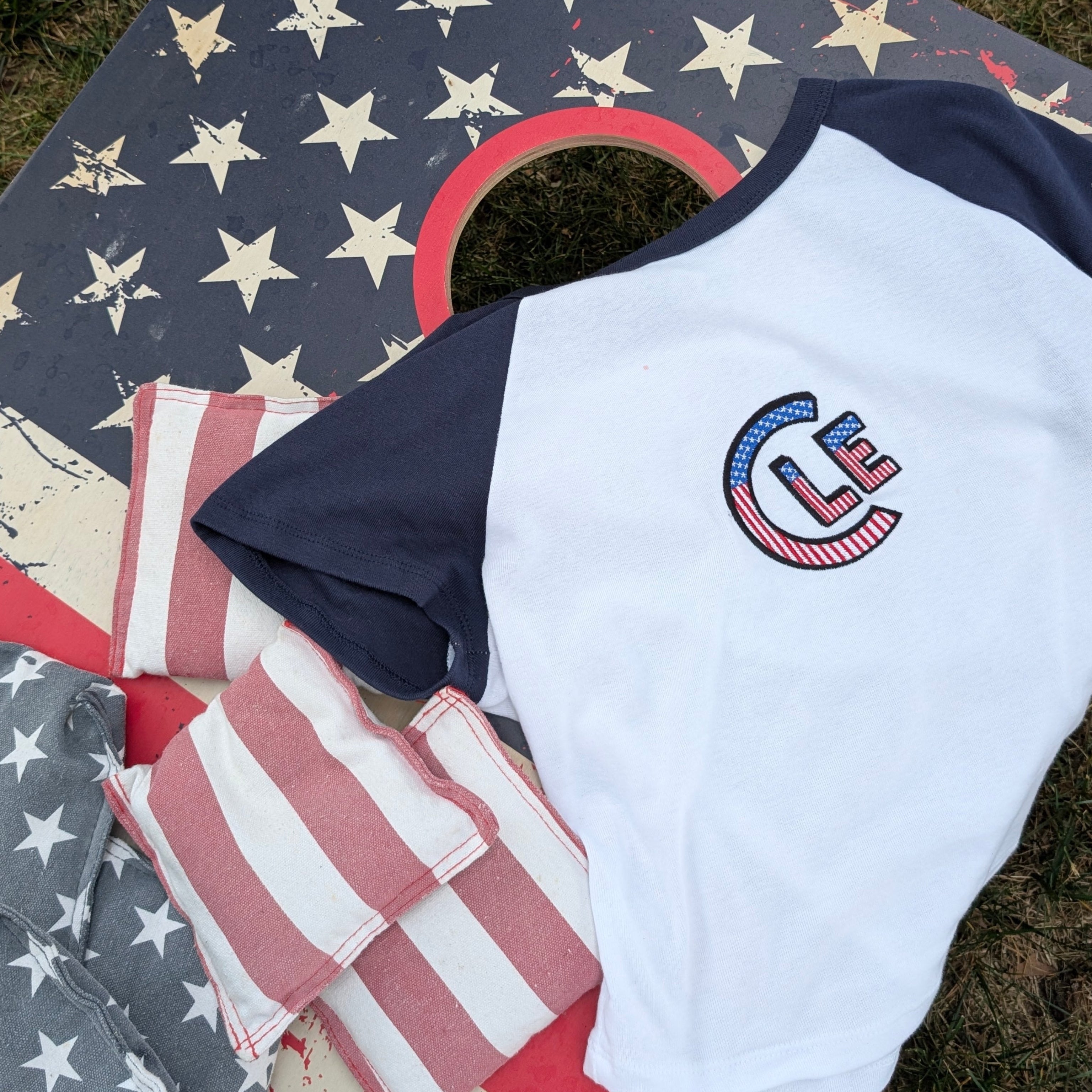 A white and navy raglan t-shirt with a patriotic 'CLE' USA graphic on the chest, laid out on a cornhole board decorated with an American flag design. The shirt is styled with matching cornhole bags on an outdoor grassy lawn.