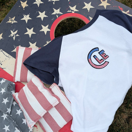 A white and navy raglan t-shirt with a patriotic 'CLE' USA graphic on the chest, laid out on a cornhole board decorated with an American flag design. The shirt is styled with matching cornhole bags on an outdoor grassy lawn.